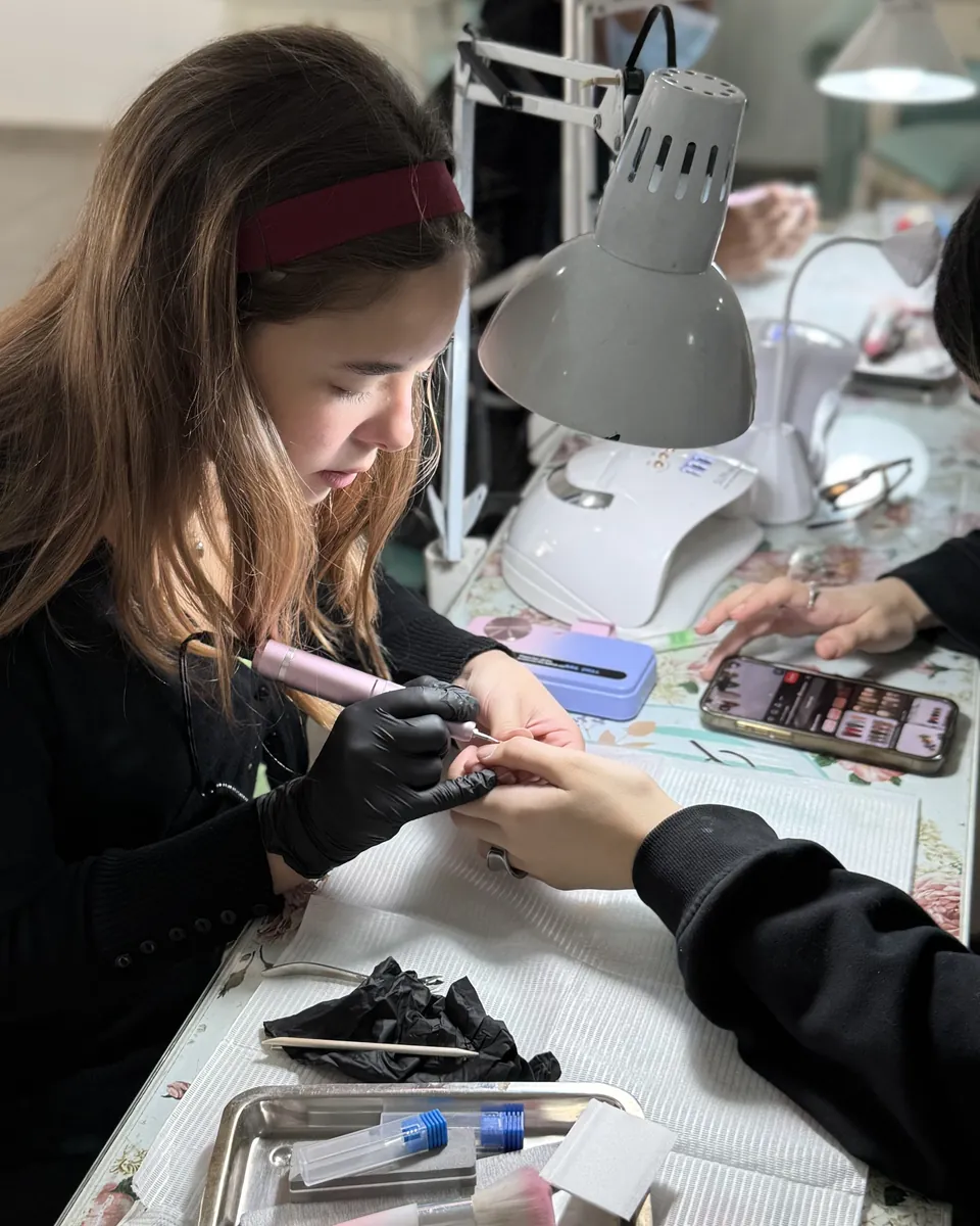 Una alumna con guantes negros trabaja con el torno sobre la mano de una modelo, en su puesto con la lámpara encendida.