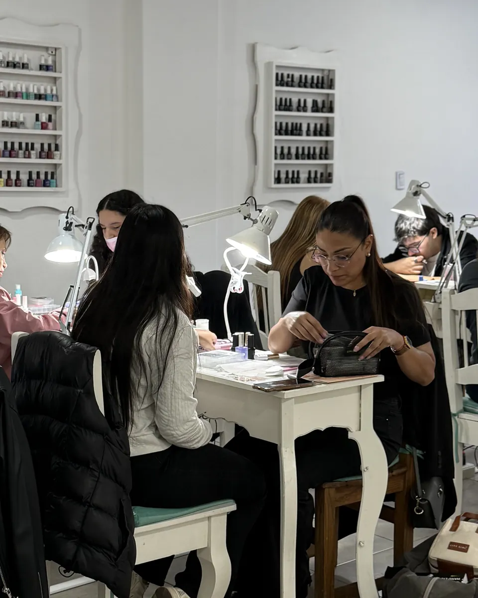 Dos alumnas trabajando en sus puestos, con los estantes de esmaltes de la escuela al fondo.