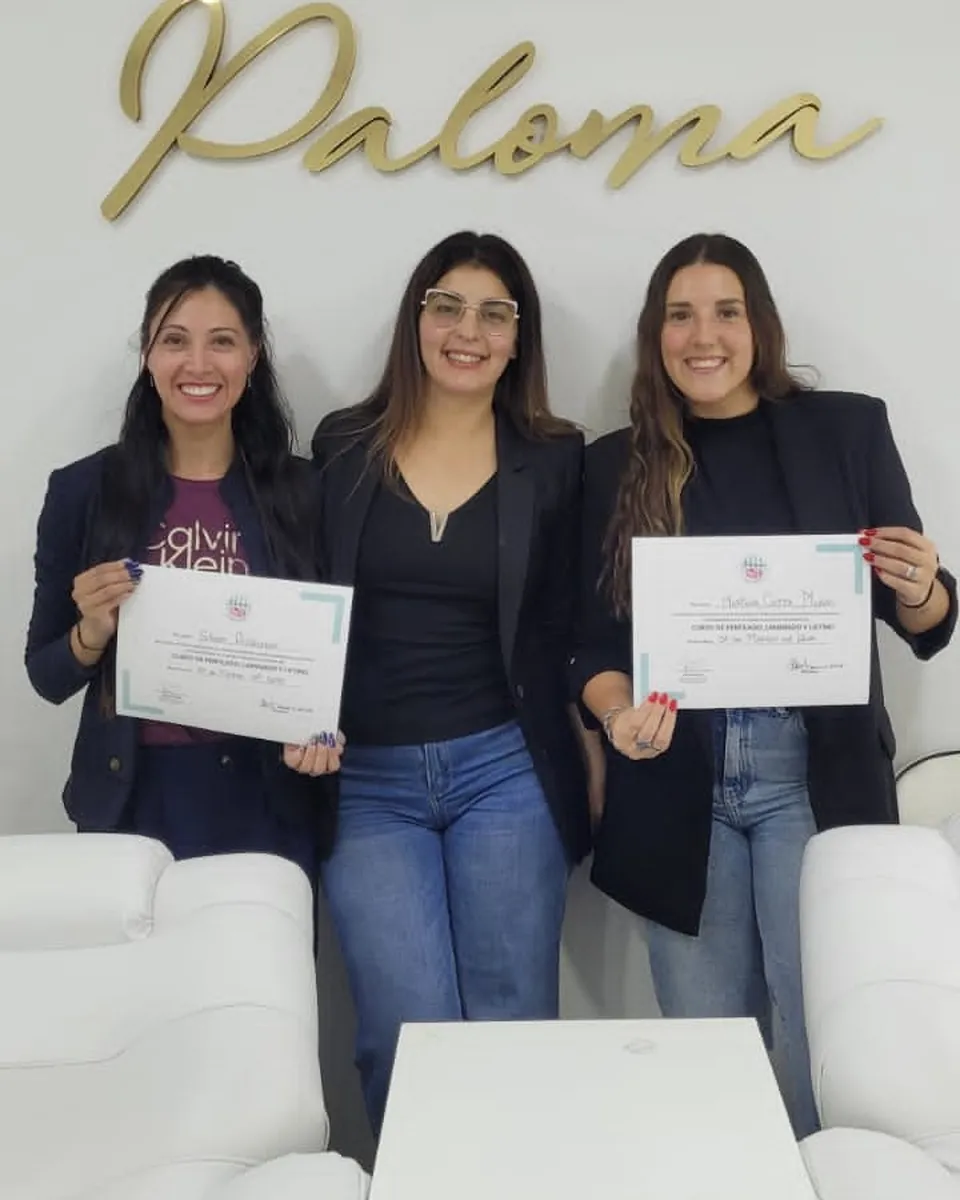Dos alumnas muestran sus certificados del curso de perfilado, laminado y lifting junto a la profesora, bajo el logo dorado de Paloma.