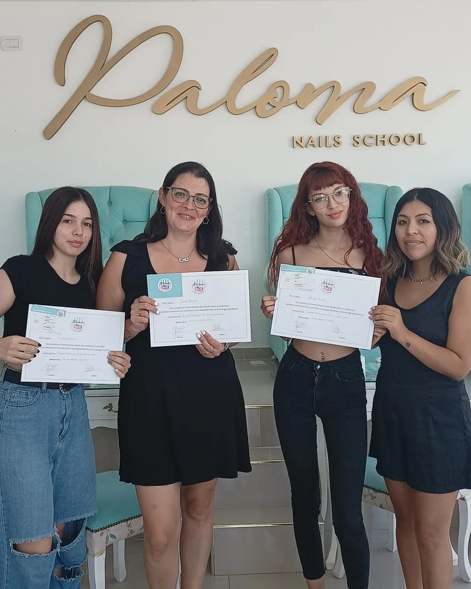 Cuatro alumnas de la Carrera Profesional de Técnica en Uñas con sus certificados, delante de los sillones turquesa de la escuela.